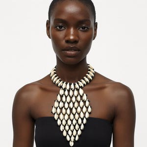 May include: A statement necklace featuring a wide collar design composed of numerous off-white cowrie shells arranged in a grid pattern, interspersed with small black beads. The shells are linked together to form a large, pointed, bib-style shape.