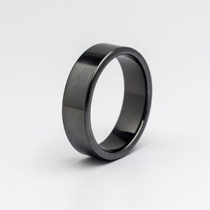 Op de afbeelding: Een zwarte ring met een platte bovenkant en een gepolijste afwerking. De ring is gemaakt van een donker, mogelijk metalen materiaal en heeft een glad, reflecterend oppervlak. De ring is een eenvoudig bandontwerp.