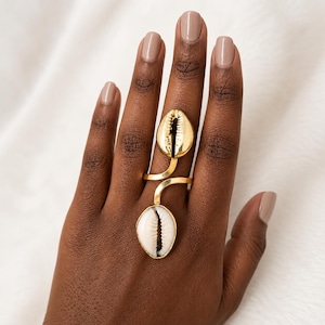 May include: A gold-toned ring featuring two cowrie shells. One shell is metallic gold and positioned near the knuckle, while the other is a natural white shell located lower on the finger. The shells are connected by a thin, curved gold metal band.