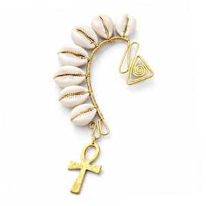 May include: A gold-toned ear cuff featuring a curved wire design adorned with six white cowrie shells. The cuff includes a geometric spiral and a hammered Ankh pendant, creating a unique and cultural accessory.