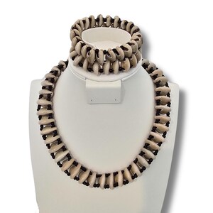 May include: A set of shell jewellery, comprising a necklace and bracelet. The necklace and bracelet are made from natural cowrie shells and black beads. The shells are a light beige colour.