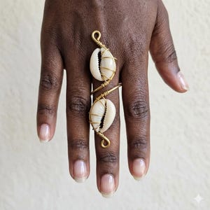 May include: A long, vertical ring featuring two white cowrie shells wrapped in gold-toned wire. The wire design extends along the length of the finger, securing each shell in place with coiled metal accents at the top, middle, and bottom ends.