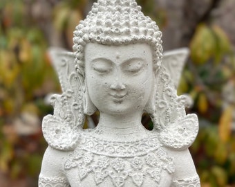 May include: A detailed, light gray statue of a serene Buddha figure with hands clasped in prayer. The statue features intricate details in the hair, clothing, and decorative elements. The background is blurred, with hints of green and yellow foliage.