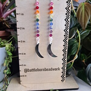 May include: Rainbow crystal beaded earrings with silver crescent moon charms. The earrings are approximately 6 inches long. The image includes the text "@battlebornbeadwork".