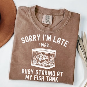 Może przedstawiać: Brązowa koszulka z białym napisem "SORRY I'M LATE I WAS... BUSY STARING AT MY FISH TANK". Grafika przedstawia akwarium z rybą i roślinami wodnymi. Koszulka ma okrągły dekolt i kr&oacute;tkie rękawy.