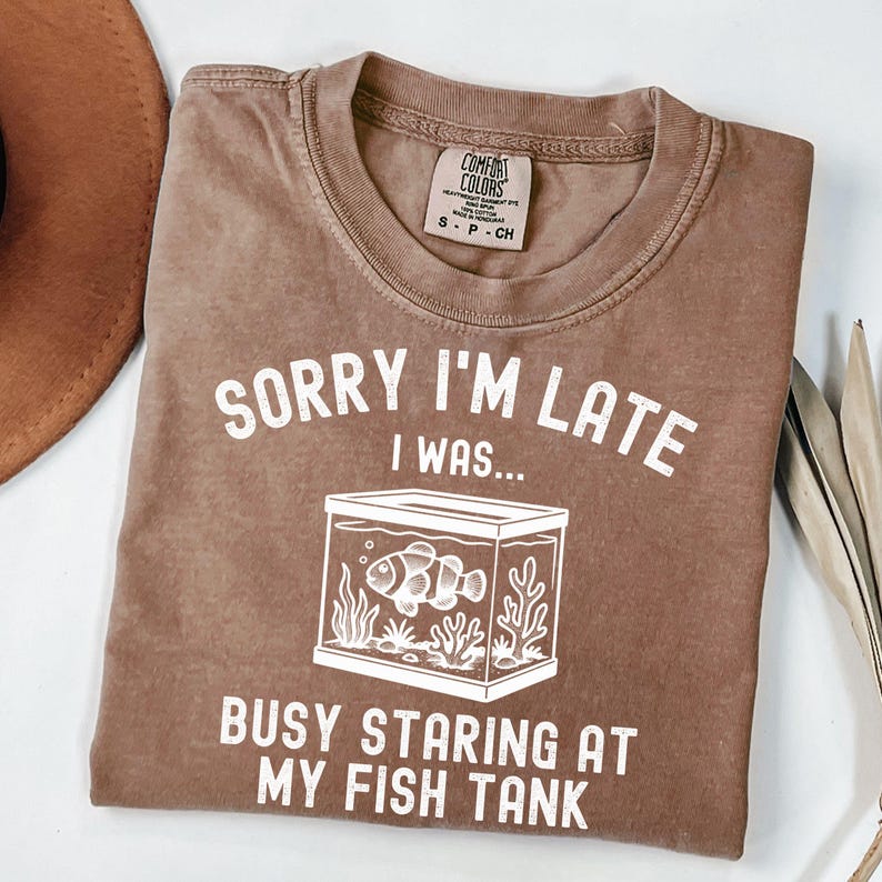 Może przedstawiać: Brązowa koszulka z białym napisem "SORRY I'M LATE I WAS... BUSY STARING AT MY FISH TANK". Grafika przedstawia akwarium z rybą i roślinami wodnymi. Koszulka ma okrągły dekolt i kr&oacute;tkie rękawy.