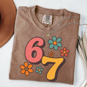 May include: Brown t-shirt with the numbers "67" in retro font, surrounded by colorful flower graphics. The shirt has a crew neck and a small tag on the upper chest. A silver necklace with a pendant is draped across the shirt.