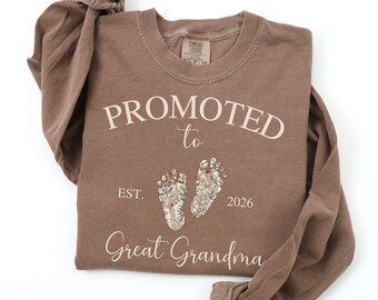 May include: A brown long-sleeved shirt with the words "PROMOTED to Great Grandma" printed in a cream color. The shirt also features two floral foot prints and the year "2026". The shirt is made of a soft material.