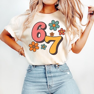 May include: Cream-colored t-shirt featuring the numbers "6" and "7" in a retro font, with colorful flower graphics. The number "6" is pink, and the number "7" is orange. The flowers are orange, teal, and pink.