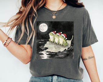 May include: Dark grey t-shirt featuring a whimsical illustration of three green frogs wearing Santa hats, floating on a leaf under a full moon. The artwork is set against a dark, watery background, creating a festive and unique design.