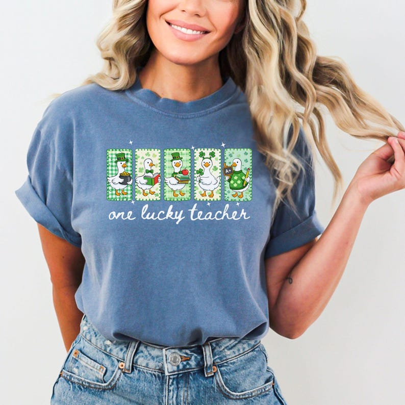 May include: A blue t-shirt with a graphic of five ducks in St. Patrick's Day attire. Each duck is in a different pose, with a green and white background. The text "one lucky teacher" is printed below the ducks.