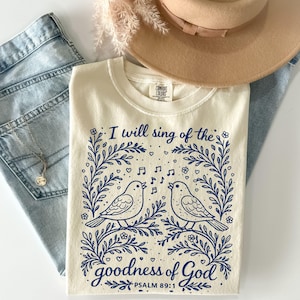 May include: A cream-coloured t-shirt with a blue floral design and the text "I will sing of the goodness of God" and "PSALM 89:1". The design features two birds, musical notes, and floral elements. The shirt is paired with blue jeans and a tan hat.