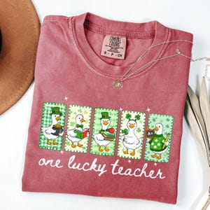May include: A maroon Comfort Colors t-shirt featuring a graphic of five ducks in St. Patrick's Day attire. Each duck holds items like a pot of gold, books, and a pencil. The text "one lucky teacher" is printed below the graphic.