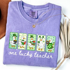 May include: A lavender Comfort Colors t-shirt featuring a graphic of five cartoon ducks, each in a St. Patrick's Day theme. The ducks are in green frames, and the text "one lucky teacher" is printed below the ducks. A gold necklace is draped across the shirt.