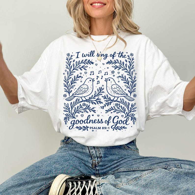 May include: White t-shirt featuring a blue illustration of two birds singing, framed by floral elements. The text "I will sing of the goodness of God" is displayed. Psalm 89:1 is also printed on the shirt.