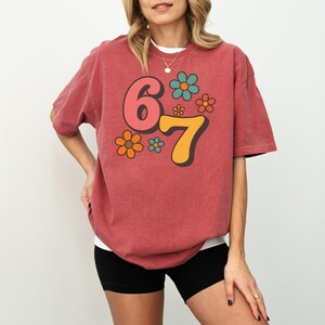 May include: A brick red t-shirt with the number "67" in a retro font, accompanied by colorful flower graphics. The shirt is a relaxed fit and has short sleeves. The model is wearing black shorts.