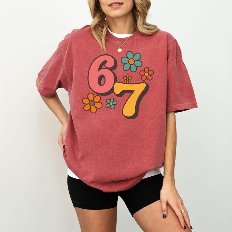May include: A brick red t-shirt with the number "67" in a retro font, accompanied by colorful flower graphics. The shirt is a relaxed fit and has short sleeves. The model is wearing black shorts.