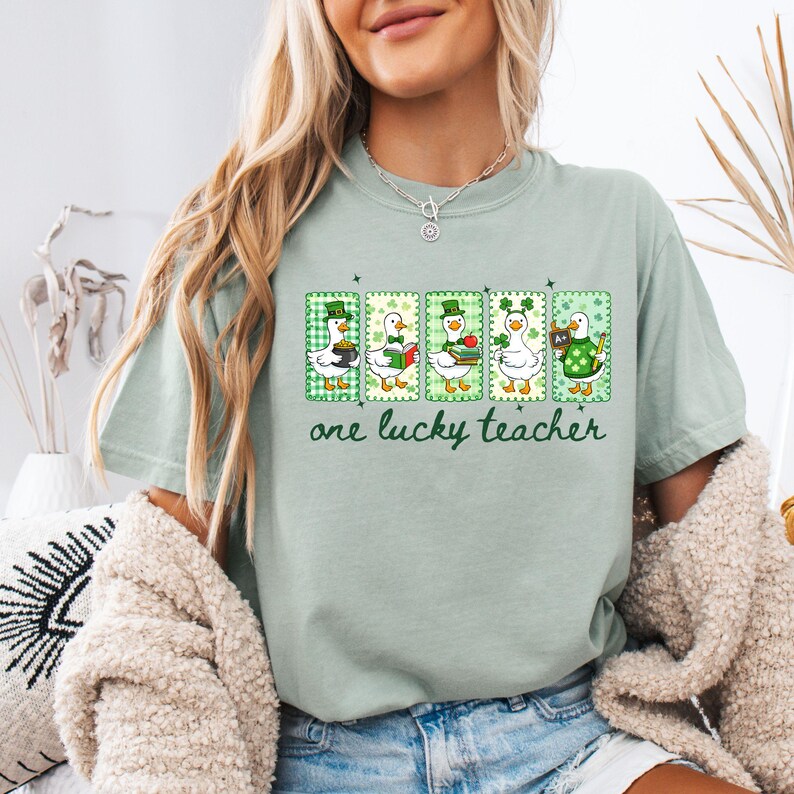 May include: A light green t-shirt featuring five cartoon ducks in St. Patrick's Day attire. Each duck is in a green frame with a shamrock design. The text "one lucky teacher" is printed in green below the ducks.