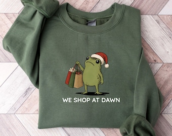 May include: A military green crewneck sweatshirt featuring a graphic of a green frog wearing a red and white Santa hat, holding three shopping bags. Below the frog, the text reads WE SHOP AT DAWN in white capital letters.