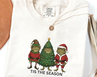 May include: Cream-colored t-shirt featuring a festive design with three frog characters dressed in Christmas attire. The frogs are depicted as an elf, a Christmas tree, and Santa Claus. The text "TIS THE SEASON" is printed below the characters.
