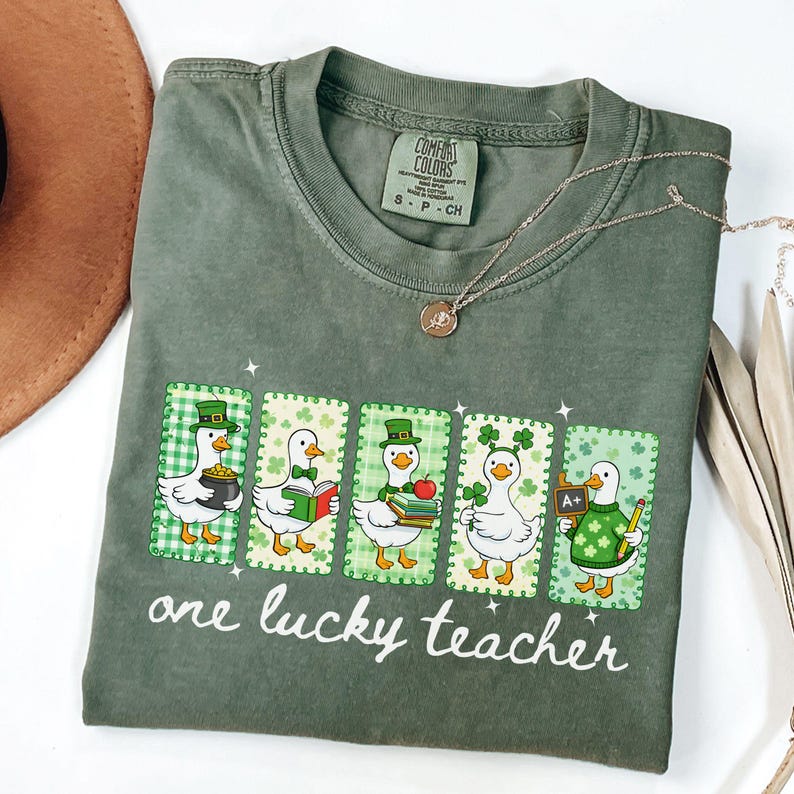 May include: Green Comfort Colors t-shirt with a graphic of five cartoon ducks in St. Patrick's Day attire. Each duck is in a different pose, holding items like a pot of gold, books, and a chalkboard. The text "one lucky teacher" is printed below.