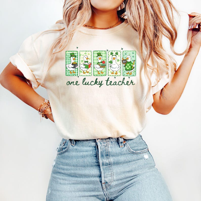 May include: Cream-colored t-shirt featuring a St. Patrick's Day design. The graphic includes four cartoon geese dressed in green and white, each holding a different item. The text "one lucky teacher" is printed below the geese.