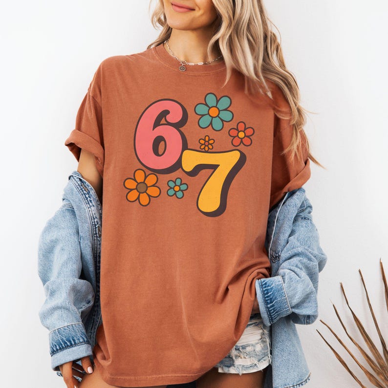 May include: A rust-colored t-shirt featuring the numbers "67" in a retro font, with a pink "6" and a yellow "7". The shirt is adorned with colorful flower graphics in shades of orange, blue, and pink. The shirt has rolled-up sleeves.