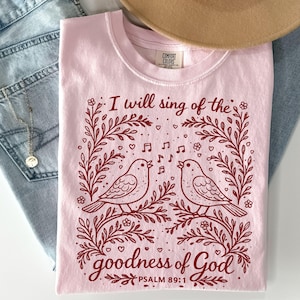 May include: A light pink Comfort Colors t-shirt with a maroon illustration of two birds singing on a branch, framed by floral elements. The text "I will sing of the goodness of God" is above the birds, with "Psalm 89:1" below.