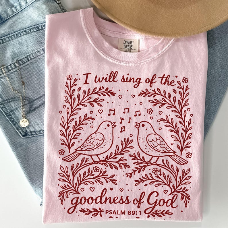 May include: A light pink Comfort Colors t-shirt with a maroon illustration of two birds singing on a branch, framed by floral elements. The text "I will sing of the goodness of God" is above the birds, with "Psalm 89:1" below.