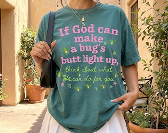 May include: A teal short-sleeve t-shirt featuring the pink and light green text If God can make a bug's butt light up, think about what He can do for you, surrounded by small yellow firefly illustrations and a black shoulder bag.