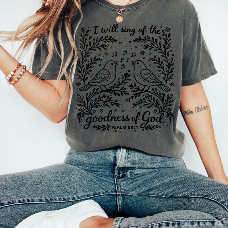 May include: Dark grey t-shirt with a black graphic of two singing birds, framed by floral designs and musical notes. The text says "I will sing of the goodness of God" and "Psalm 89:1".