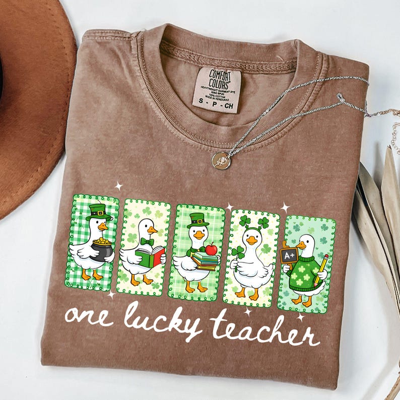 May include: Brown t-shirt with a graphic of five cartoon ducks in St. Patrick's Day attire. Each duck is in a different pose, holding a pot of gold, a book, an apple, a shamrock, and a chalkboard. The text "one lucky teacher" is below the graphic.