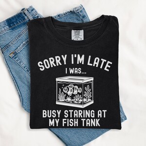 Może przedstawiać: Czarny t-shirt z białym napisem "SORRY I'M LATE I WAS... BUSY STARING AT MY FISH TANK". Grafika zawiera akwarium z błazenkiem. Koszulka jest złożona na niebieskich dżinsach.