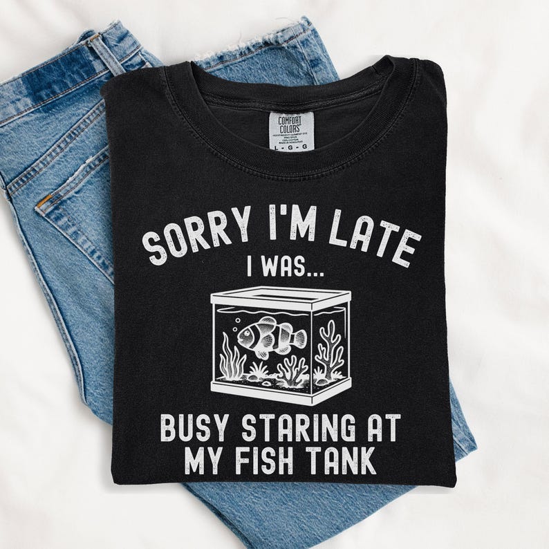 Może przedstawiać: Czarny t-shirt z białym napisem "SORRY I'M LATE I WAS... BUSY STARING AT MY FISH TANK". Grafika zawiera akwarium z błazenkiem. Koszulka jest złożona na niebieskich dżinsach.