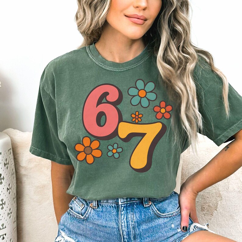 May include: Green t-shirt with the number "67" in pink and yellow, surrounded by colorful flowers. The shirt has a vintage, retro design, perfect for a casual, everyday look.