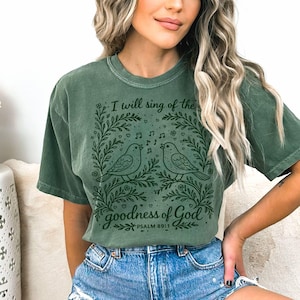 May include: Green t-shirt with a graphic of two singing birds within a floral wreath. The text reads "I will sing of the goodness of God" above the birds, with "Psalm 89:1" below. The shirt is a muted green colour.