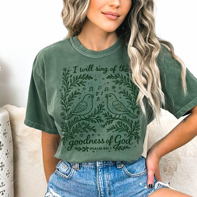 May include: Green t-shirt with a graphic of two singing birds within a floral wreath. The text reads "I will sing of the goodness of God" above the birds, with "Psalm 89:1" below. The shirt is a muted green colour.
