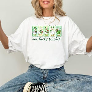 May include: White t-shirt with a green and white graphic featuring five cartoon ducks in St. Patrick's Day attire. The text "one lucky teacher" is printed below the ducks in green script. The shirt is made of a soft material.