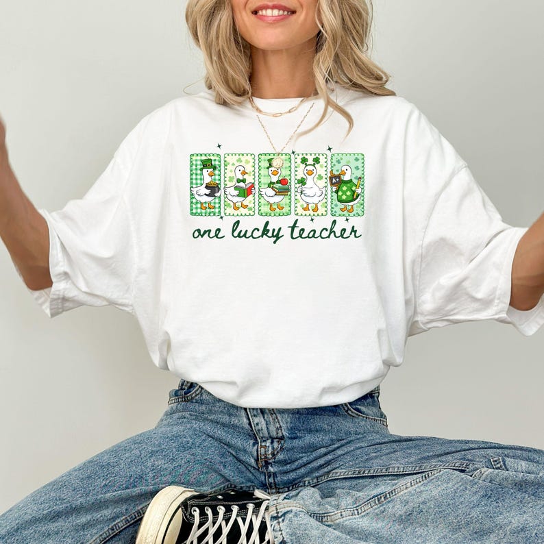 May include: White t-shirt with a green and white graphic featuring five cartoon ducks in St. Patrick's Day attire. The text "one lucky teacher" is printed below the ducks in green script. The shirt is made of a soft material.