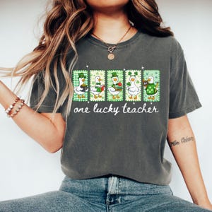 May include: Dark gray t-shirt featuring a graphic of five ducks in St. Patrick's Day attire. Each duck is in a different pose, holding items like a mug, book, and apple. The text "one lucky teacher" is below the ducks.