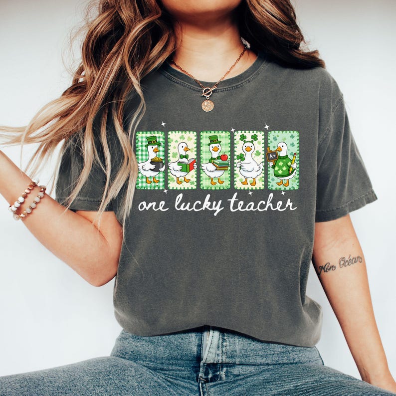 May include: Dark gray t-shirt featuring a graphic of five ducks in St. Patrick's Day attire. Each duck is in a different pose, holding items like a mug, book, and apple. The text "one lucky teacher" is below the ducks.