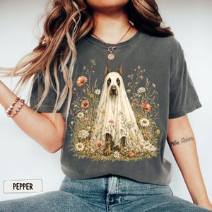 May include: A charcoal gray t-shirt featuring a graphic print of a Doberman dog draped in a white translucent sheet, resembling a ghost, surrounded by a colorful array of wildflowers. The shirt has a crew neckline and short sleeves.