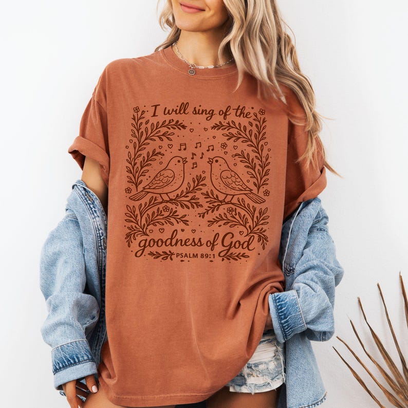 May include: A terracotta-coloured t-shirt with a brown graphic of two birds singing, framed by floral designs and musical notes. The text says "I will sing of the goodness of God" and "Psalm 89:1".