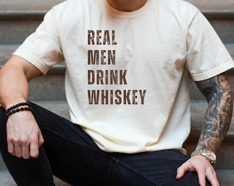 May include: A beige t-shirt with the text "REAL MEN DRINK WHISKEY" printed in brown on the front.