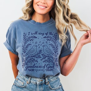 May include: A blue t-shirt with a graphic design featuring two birds singing, surrounded by floral elements and musical notes. The text reads "I will sing of the goodness of God" and "Psalm 89:1".