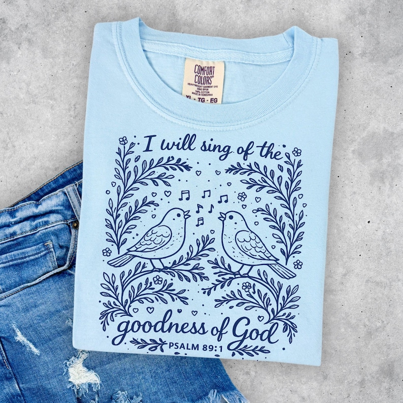 May include: Pale blue t-shirt with a graphic of two singing birds on a branch, framed by floral designs and musical notes. The text reads "I will sing of the goodness of God, Psalm 89:1". The shirt has a Comfort Colors label.