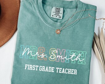 May include: A teal Comfort Colors t-shirt with the text "Mrs. Smith First Grade Teacher" in colourful block letters and white script. A gold necklace with a pendant rests on the shirt.