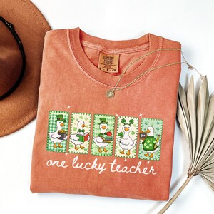 May include: Coral-colored t-shirt featuring a graphic of five cartoon ducks dressed for St. Patrick's Day. Each duck is in a green and white checkered frame. The text "one lucky teacher" is printed below the graphic.