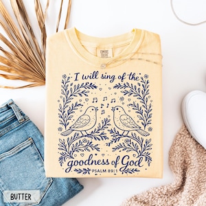 May include: A pale yellow sweatshirt with a navy blue graphic of two singing birds, musical notes, and the text "I will sing of the goodness of God" from Psalm 89:1. The sweatshirt is folded and displayed with a pair of jeans and a white shoe.