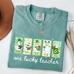 May include: A seafoam green t-shirt featuring a graphic of five cartoon ducks in St. Patrick's Day attire. Each duck is in a framed design with a green and white checkered background. The text "one lucky teacher" is printed below the ducks.